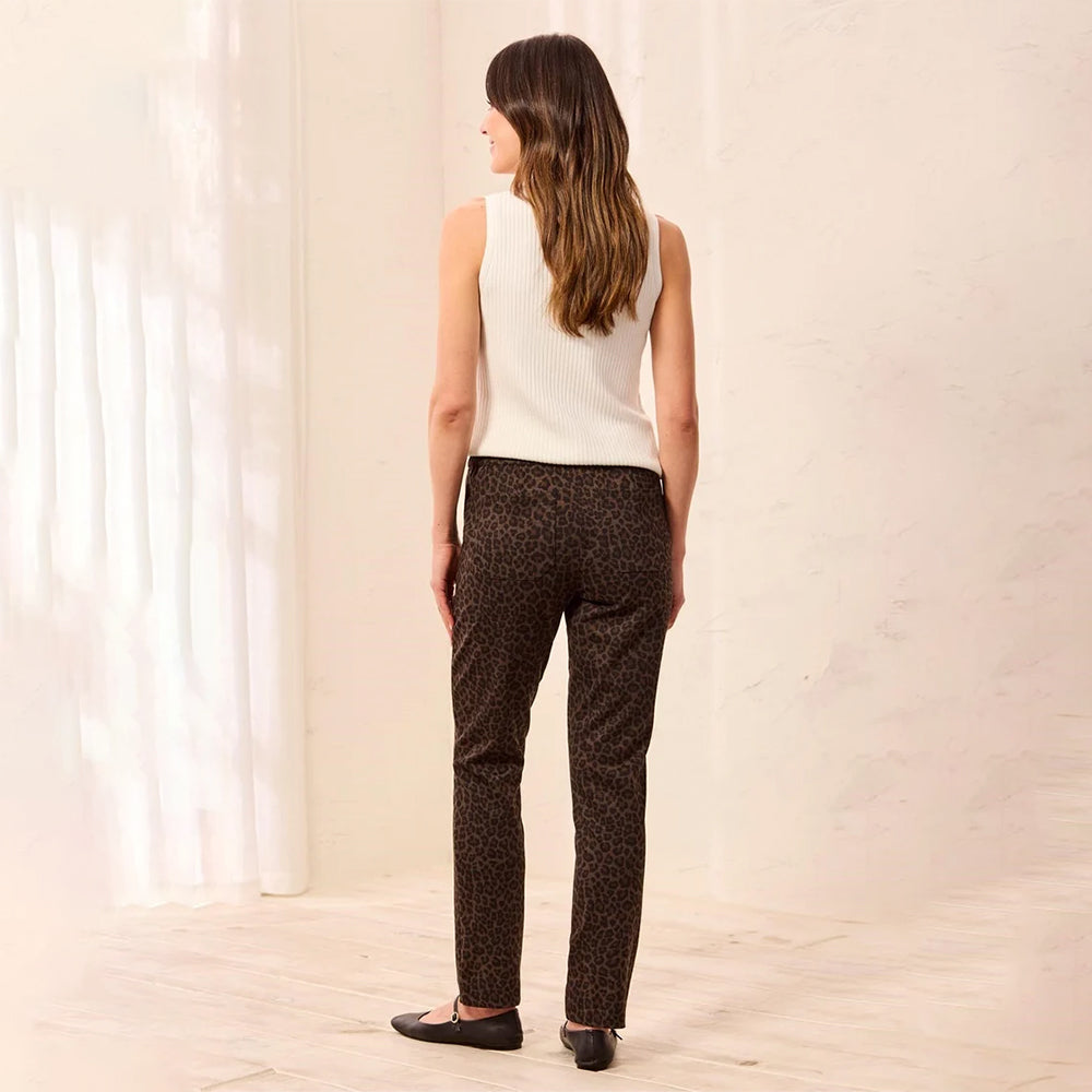 A woman stands with her back to the camera wearing dark brown leopard-print pants, a white sleeveless knit top, and black flats in a bright, minimalist room with white walls and curtains.