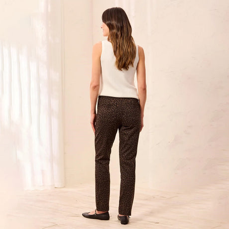 A woman stands with her back to the camera wearing dark brown leopard-print pants, a white sleeveless knit top, and black flats in a bright, minimalist room with white walls and curtains.