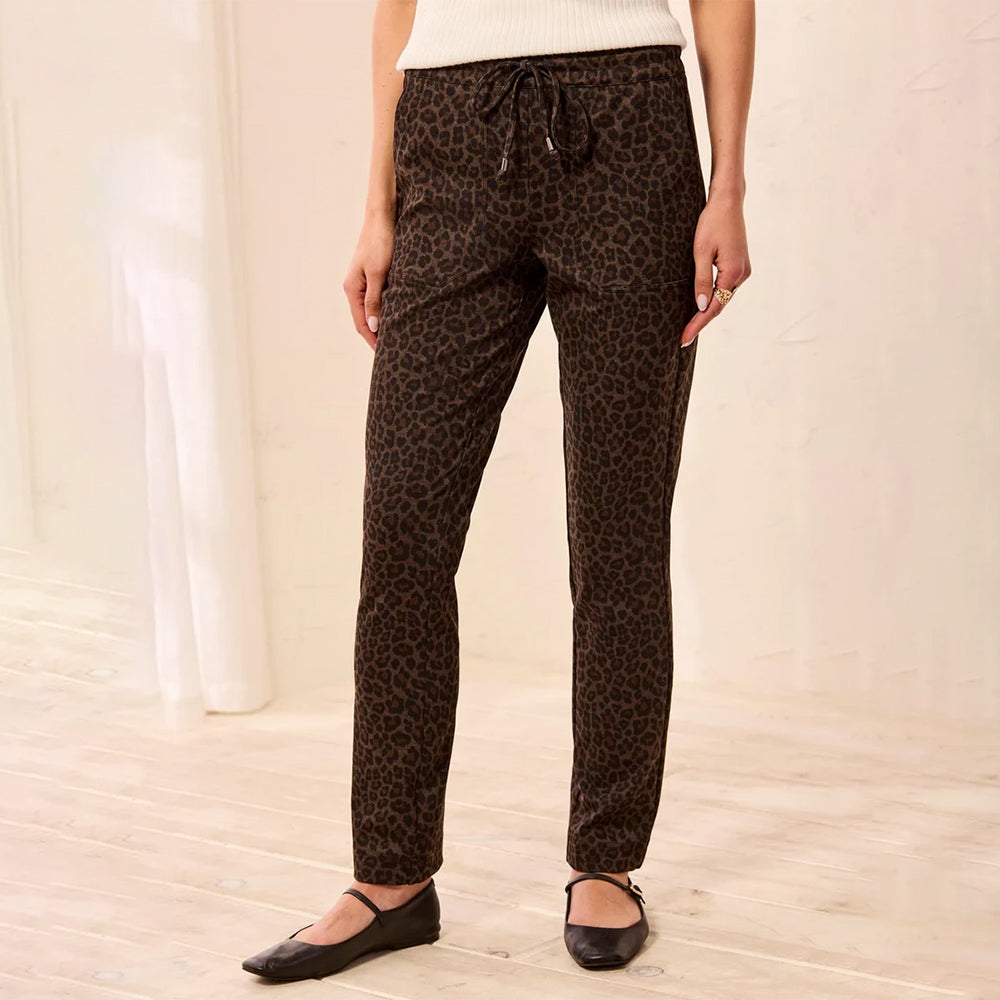 A person wearing brown leopard-print pants with a drawstring waist, paired with a white ribbed top and black flats, standing in a bright indoor room with light wood flooring.