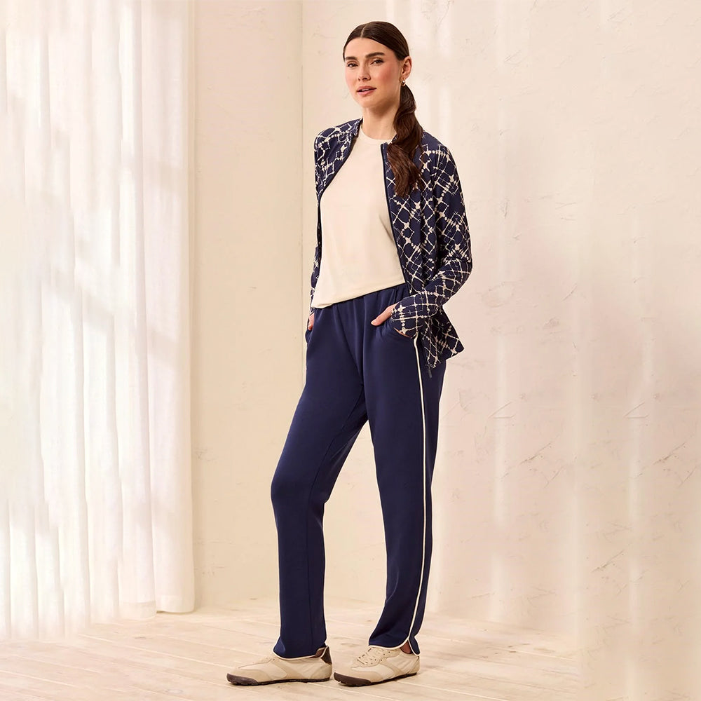 A woman stands in a softly lit indoor setting wearing navy blue track pants with a white side stripe, a cream top, and a navy-and-white patterned jacket.