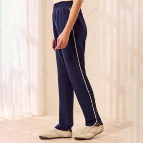A person wearing navy blue athletic pants with a white side stripe, paired with beige sneakers, standing in a brightly lit room with soft window shadows.