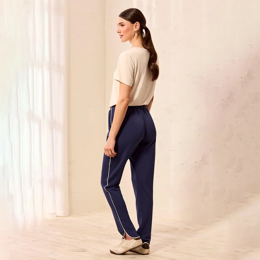 A woman stands in profile wearing navy blue athletic pants with a thin white side stripe, a light beige short-sleeve top, and beige sneakers in a bright indoor room with soft window light and curtains.
