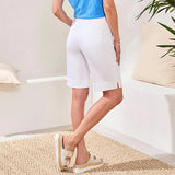 Person wearing white shorts and beige sandals in a room with a white wall and a plant.