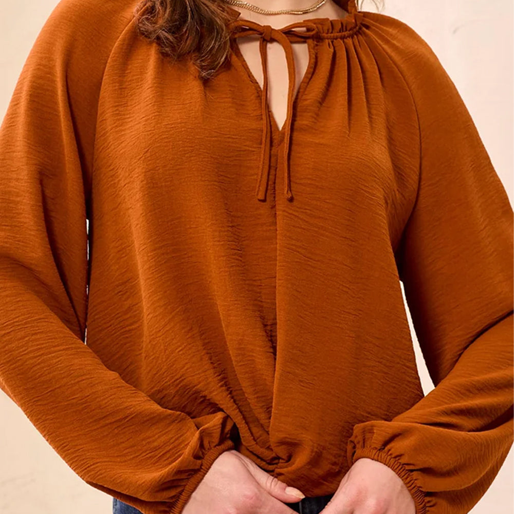 A close-up of a woman wearing a rust-orange long-sleeve blouse with a gathered keyhole neckline and tie-front detail.