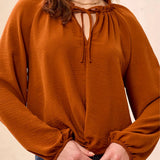 A close-up of a woman wearing a rust-orange long-sleeve blouse with a gathered keyhole neckline and tie-front detail.
