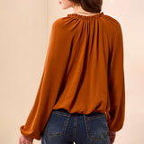 A woman wearing a rust-orange long-sleeve blouse with a gathered, pleated back neckline, shown from behind against a plain beige background.
