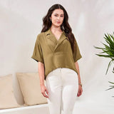 Woman wearing a green top and white pants standing in a room with a couch and plant.