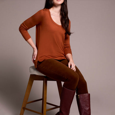 Woman wearing a rust-colored long-sleeve top and brown pants sitting on a wooden stool against a plain background.