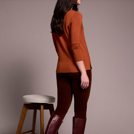 Woman wearing a rust-colored sweater and brown pants standing next to a stool against a plain background