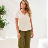 Woman wearing a white top and green pants standing in a bright room with a plant.