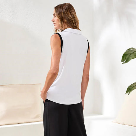 Woman wearing a white sleeveless top and black pants in a minimalistic room.
