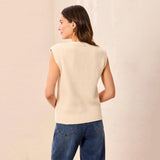A woman shown from the back wearing a sleeveless cream ribbed knit sweater vest and blue jeans, standing against a light beige textured wall.