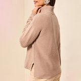 A woman wearing a ribbed knit turtleneck sweater in a light taupe-beige color, shown from a side-back angle against a soft cream background.