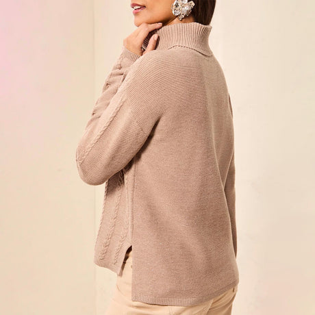 A woman wearing a ribbed knit turtleneck sweater in a light taupe-beige color, shown from a side-back angle against a soft cream background.