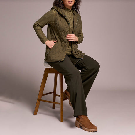 Woman wearing green zipper vest with brown pants and brown boots, sitting on a stool.