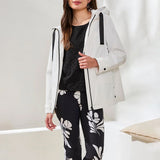 Woman wearing a white coat over a black top and floral pants in a minimal indoor setting.