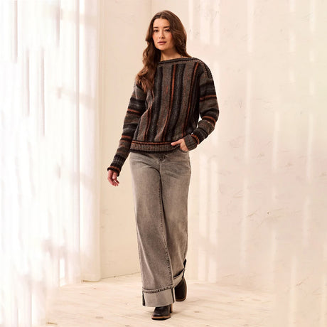 A woman modeling a dark gray and brown striped knit sweater with gray wide-leg jeans and black boots, standing in a brightly lit room with white curtains.