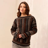 A woman wearing a dark gray and brown striped knit sweater with orange-brown vertical accents, paired with light gray jeans, standing against a neutral beige backdrop.