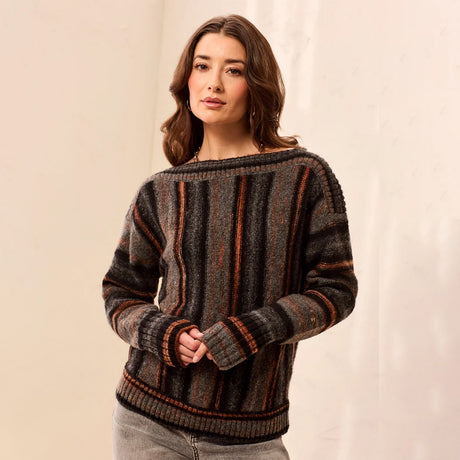 A woman wearing a dark gray and brown striped knit sweater with orange-brown vertical accents, paired with light gray jeans, standing against a neutral beige backdrop.
