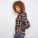 A woman with curly brown hair wearing a beige and black leopard print knit hoodie with red trim, paired with blue jeans, photographed against a plain white studio background.