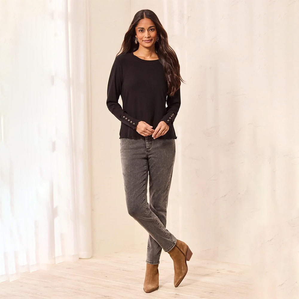 A woman modeling a black long-sleeve top with decorative silver studs on the cuffs, paired with gray jeans and brown ankle boots, standing in a bright indoor setting with a cream textured wall and light wood floor.